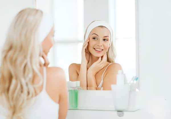 beauty, skin care and people concept - smiling young woman in hairband touching her face and looking to mirror at home bathroom